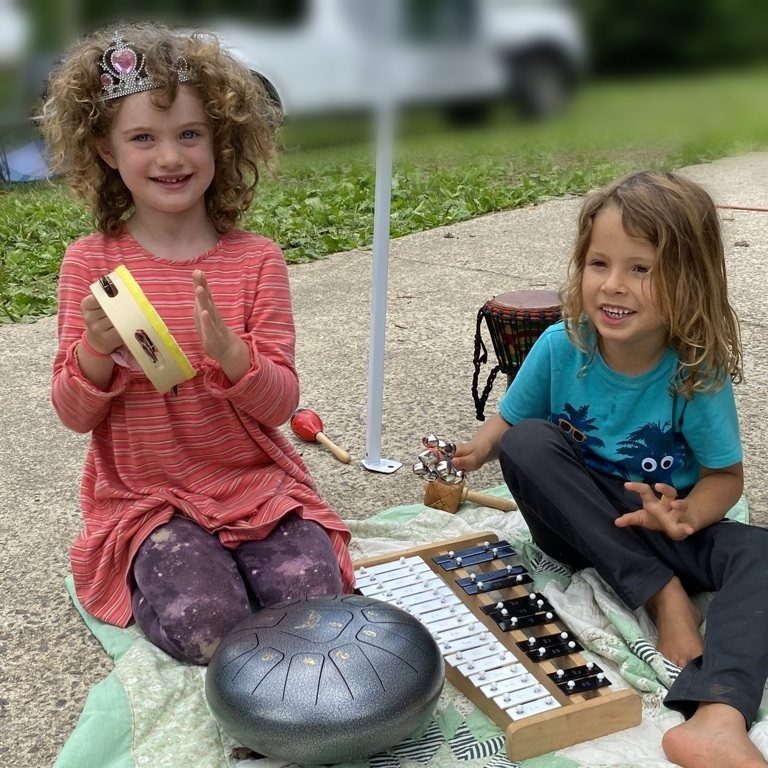 Two smiling children sitting on a quilt playing a tambourine, xylophone and drums at an outdoor market