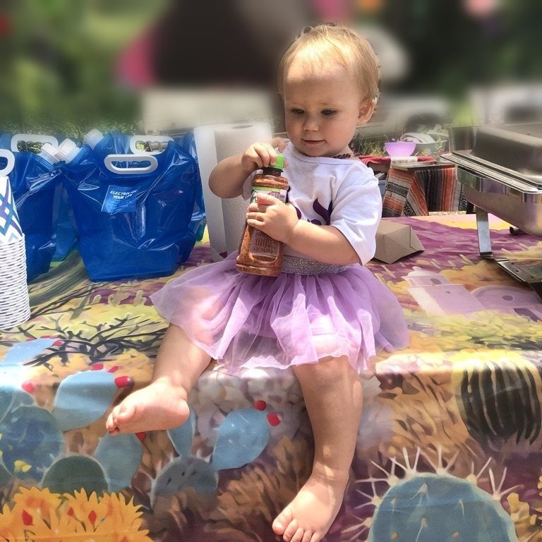 A toddler in a purple tutu sitting on a vendor's table at an outdoor market