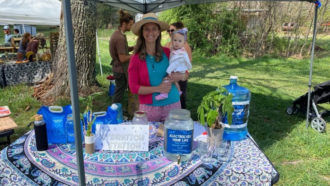 A vendor holding a baby behind her booth table under a canopy tent at an outdoor market